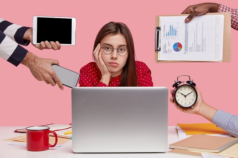 Serious Caucasian woman with dark hair, wears round spectacles, red shirt, sits in front of laptop computer drinks beverage, unrecognizable people hold alarmclock, papers, touchpad, cellular in hands
